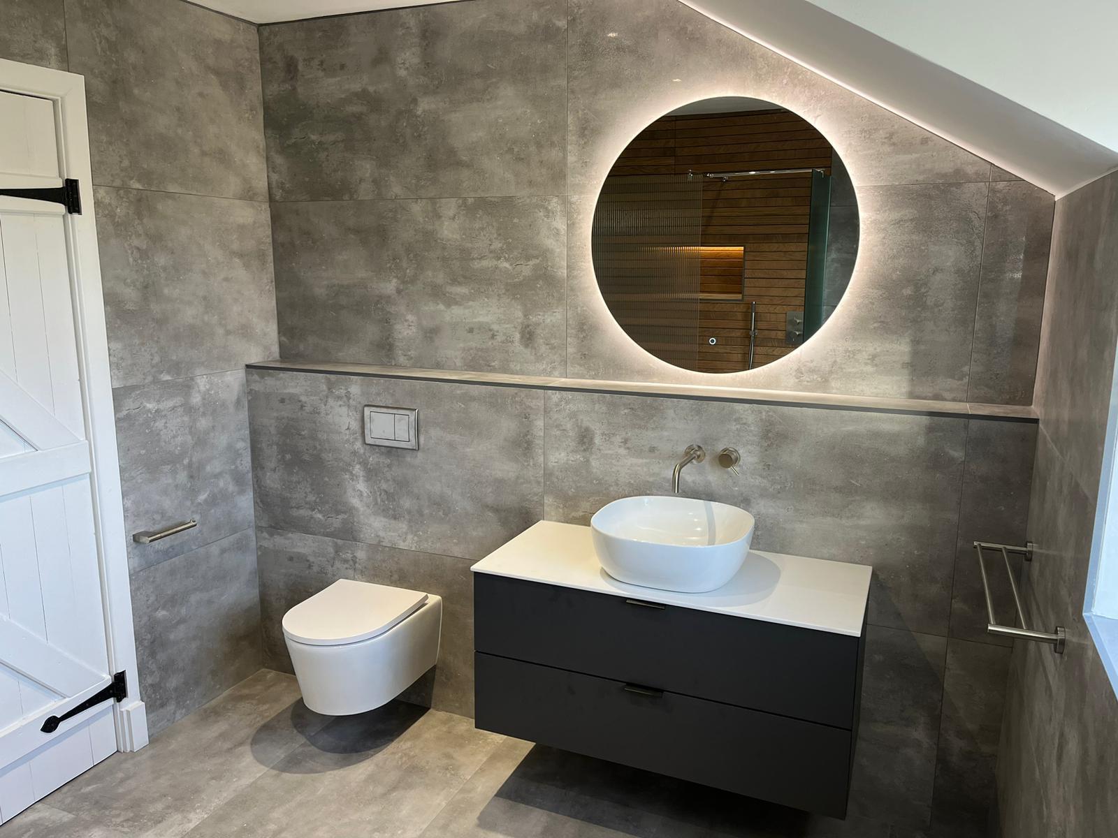 Attic ensuite with concrete-effect porcelain, backlit round mirror, graphite vanity and wall-hung WC