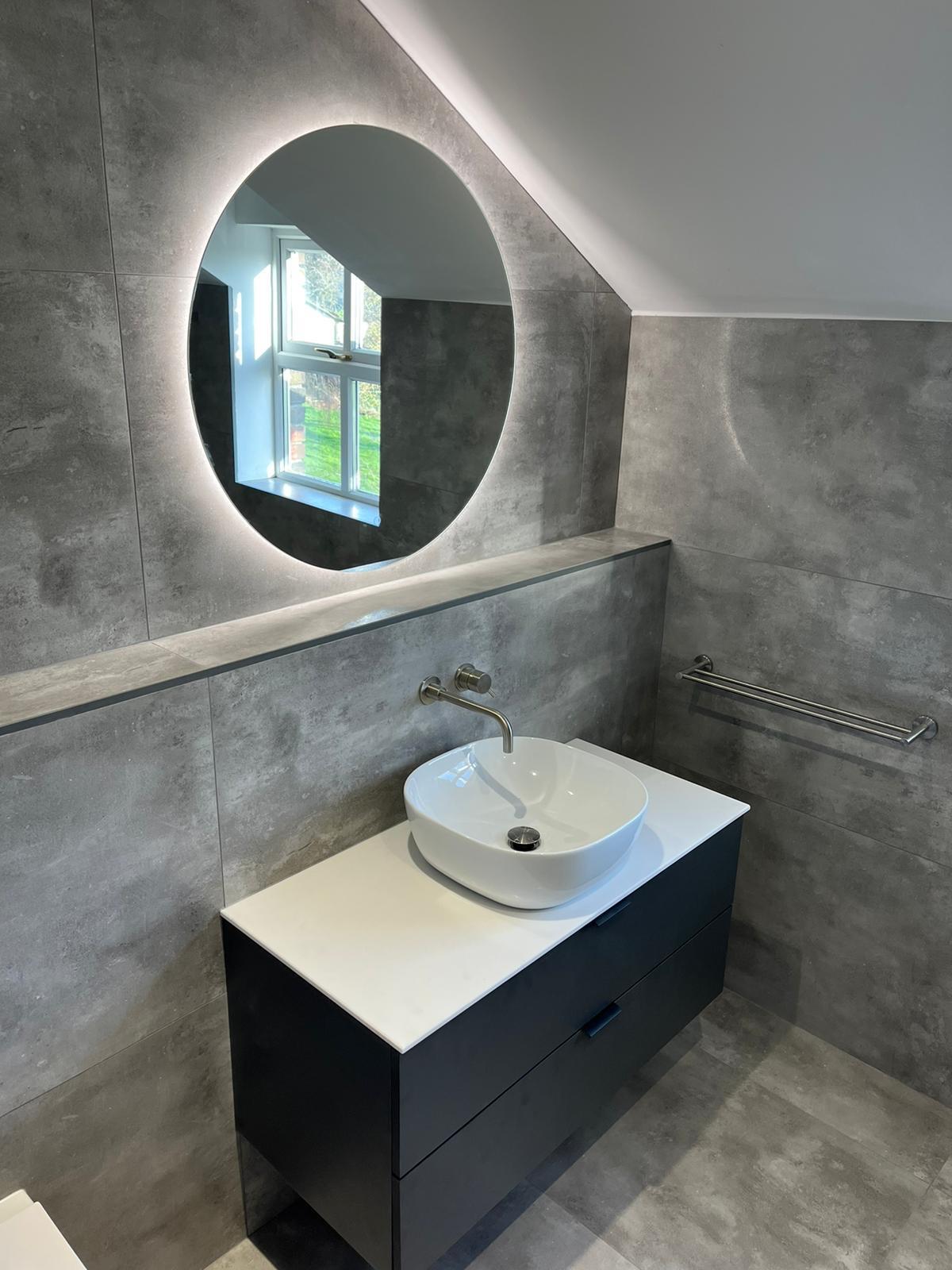 Backlit round mirror above a graphite wall-hung vanity with white quartz top and counter basin