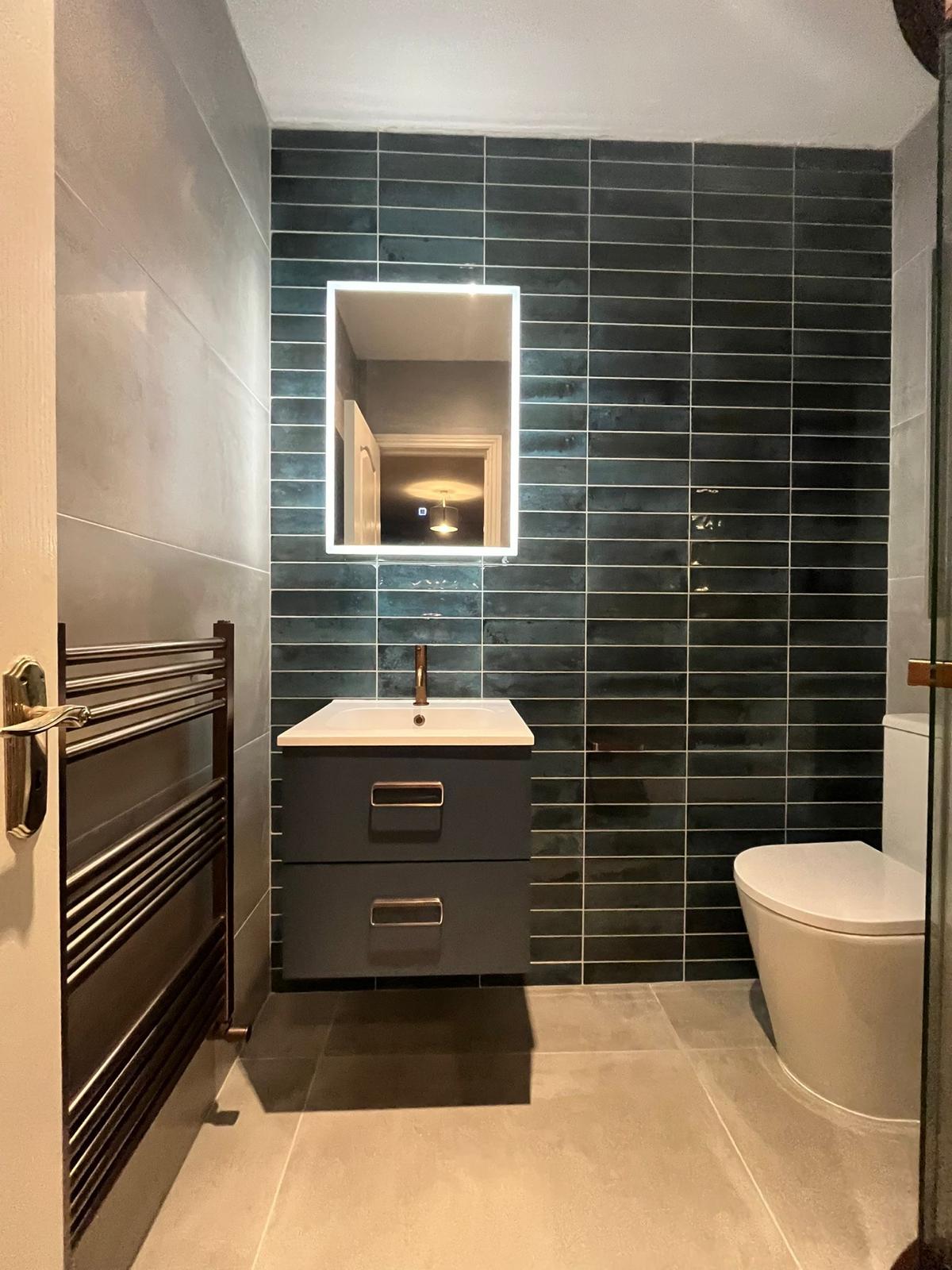 Anthracite wall-hung vanity with copper handles under a backlit mirror against teal brick-bond tiling